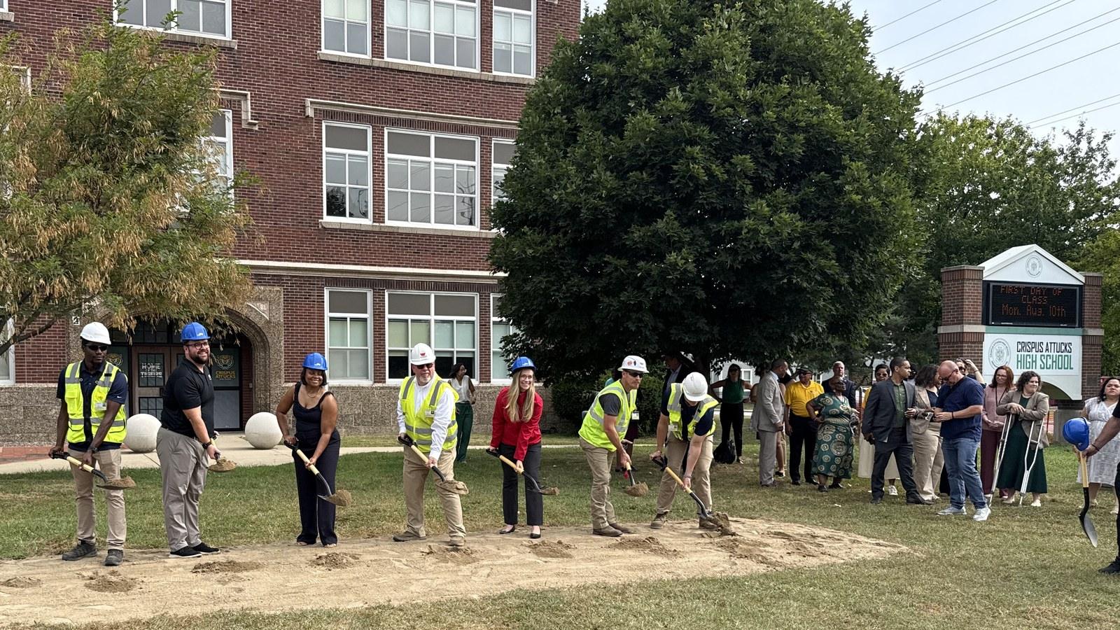 Project team members and district officials in hard hats and safety vests turn the first shovels of dirt on the lawn at Crispus Attucks High School, with the historic brick building and the school marquee behind them.