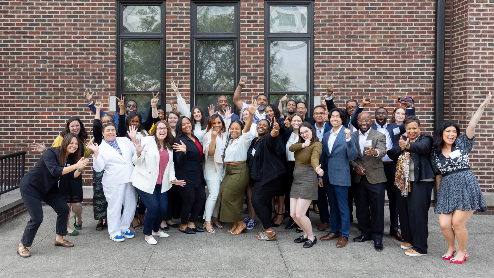 The 2025 ULI Indiana REDI cohort, about thirty graduates in business attire, cheering with raised arms outside a brick building.